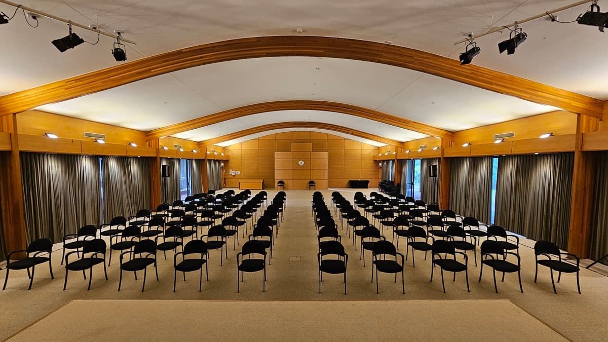 A large conference room with rows of black chairs arranged in the centre, facing a wooden panelled wall at the back. The ceiling is arched with wooden beams, and the walls are lined with long, dark curtains. The floor is carpeted in a light colour. Overhead, there are several spotlights attached to the ceiling beams. A clock is mounted on the back wall, and there are two microphones on stands at the front.