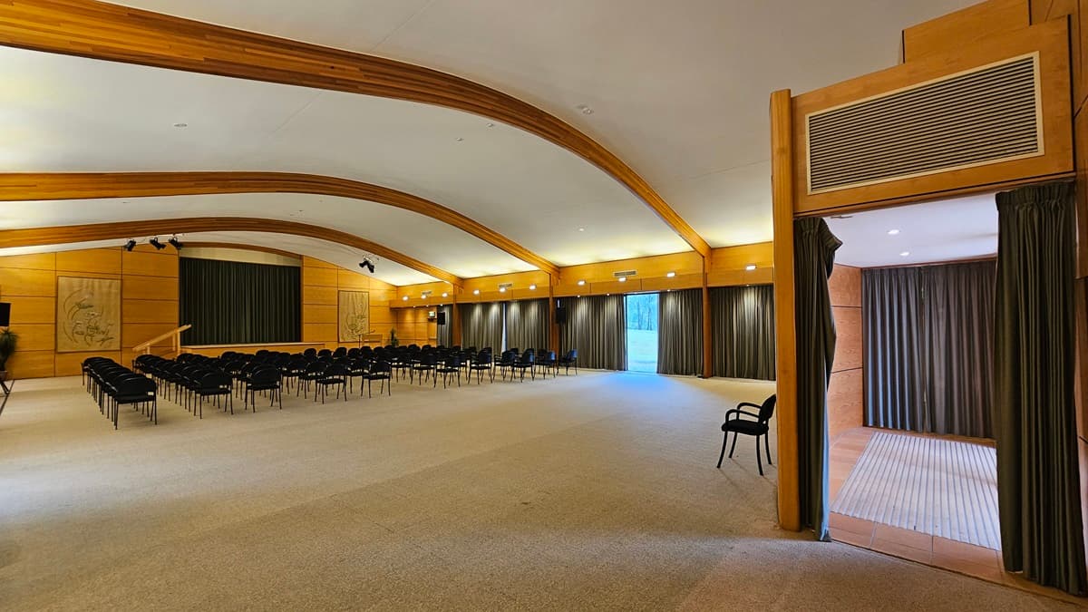A spacious conference room with a high, curved ceiling featuring wooden beams. On the left, rows of black chairs face a stage with a green curtain. The walls are wooden with decorative panels. On the right, a doorway with dark curtains leads to another room. The floor is carpeted in a light colour, and the room is well-lit with recessed lighting along the walls.