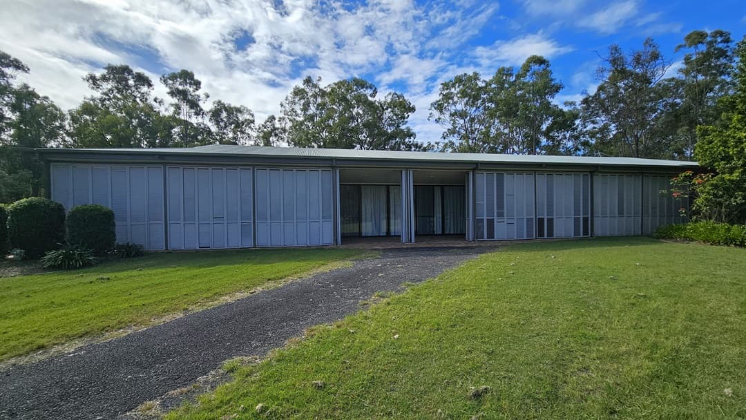A single-storey building with a flat roof and large, vertical metal panels covering the front. A gravel path leads up to the entrance, flanked by green lawns on either side. Bushes and trees are visible on the left and right, with taller trees in the background. The sky is partly cloudy.