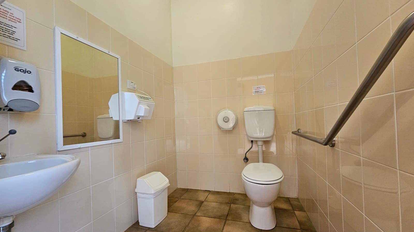A public restroom with beige tiled walls and a brown tiled floor. On the left, there's a small white sink with a soap dispenser above it and a mirror. A paper towel dispenser is mounted on the wall. In the centre, a white toilet with a wall-mounted cistern and a toilet paper holder. On the right, a metal grab bar is attached to the wall. A small white bin is on the floor near the sink. The ceiling is plain and light-coloured.