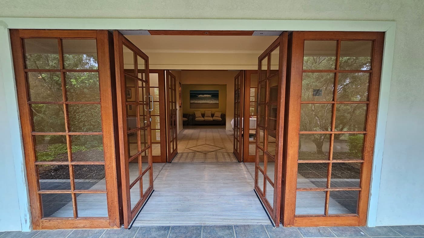 Wooden-framed glass doors open inward, leading to an interior space. The floor is tiled with a light-coloured mat at the entrance. Inside, a beige sofa with cushions is visible, set against a wall with a large painting above it. The ceiling is plain and light-coloured. On either side of the doors, there are windows showing greenery outside.