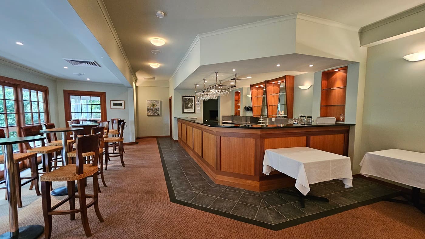 A bar area with a wooden counter on the right, featuring hanging glasses and shelves with bottles. Two tables with white tablecloths are in front of the bar. On the left, there are tall tables and chairs near large windows with wooden frames. The floor is carpeted, and the ceiling has recessed lighting. Walls are light-coloured with framed pictures.