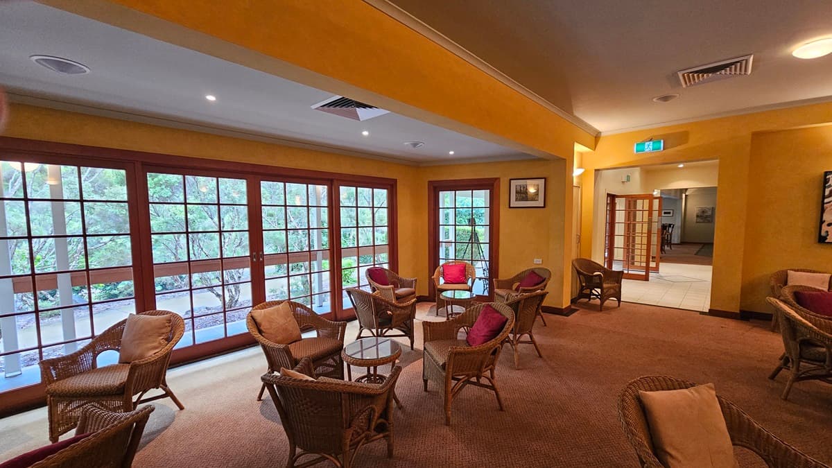 A cosy lounge area with wicker chairs and tables, each adorned with red cushions, arranged on a brown carpet. Large wooden-framed windows on the left offer a view of greenery outside. The walls are painted yellow, and the ceiling features recessed lighting and air vents. A doorway on the right leads to another room, with an exit sign above. A framed picture hangs on the wall near the doorway.