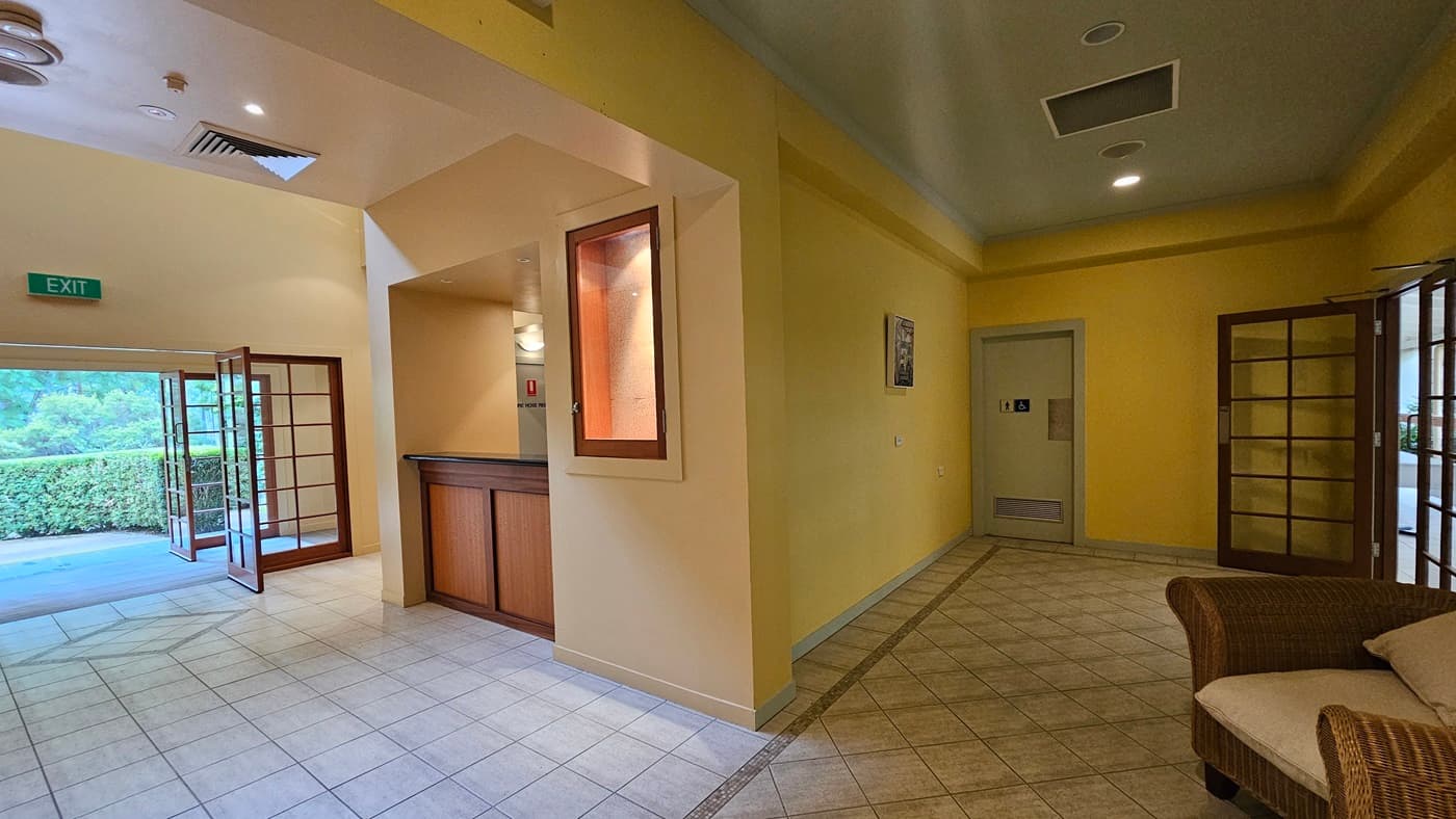 A hallway with yellow walls and a tiled floor. On the left, there is an open wooden-framed glass door leading outside, with an "EXIT" sign above. A wooden counter is visible near the centre, with a small window above it. On the right, a wicker chair with a cushion is placed against the wall. Further down the hallway, a door with a sign is visible. The ceiling has recessed lighting and ventilation panels.