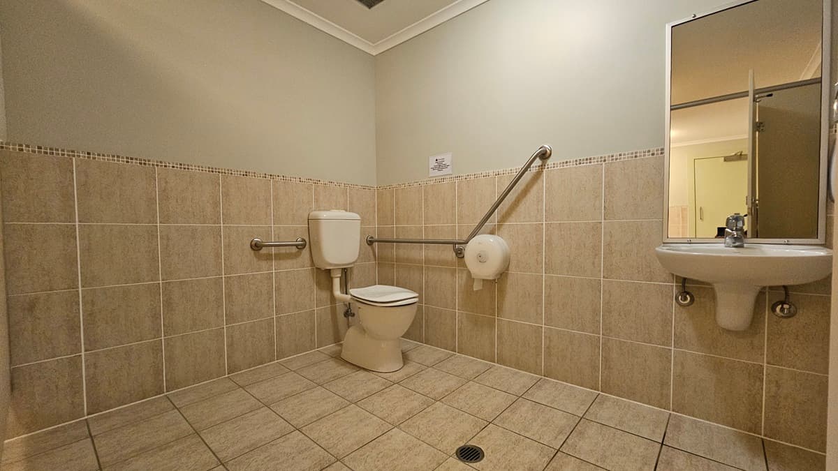A bathroom with beige tiled walls and floor. On the left, there is a toilet with a white cistern, equipped with horizontal and diagonal metal grab bars. A toilet paper holder is attached to the diagonal bar. On the right, a white sink with a mirror above it is visible. The ceiling is plain with a light fixture.