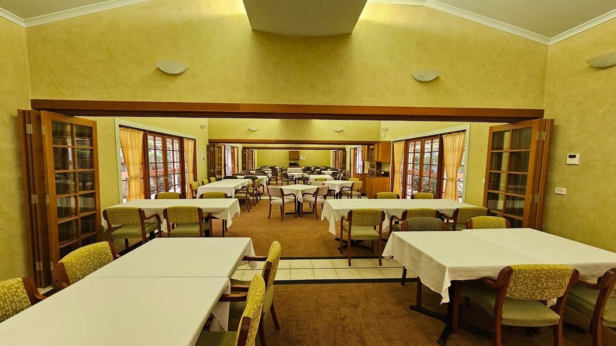 A spacious dining room with multiple rectangular tables covered in white tablecloths, surrounded by cushioned chairs. The room features large windows with wooden frames and curtains on both sides, allowing natural light to enter. The walls are painted in a warm beige tone, and there are wall-mounted lights. The floor is carpeted, and the ceiling is high with a central recessed section.