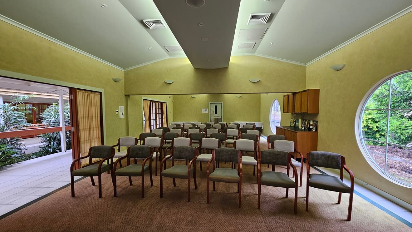 A room with rows of chairs facing forward, arranged in a semi-circle. The walls are painted yellow, and there are circular windows on the right side, offering a view of greenery outside. On the left, there is an open doorway leading to a corridor with plants visible. The ceiling is vaulted with recessed lighting and air vents. A cabinet with framed photos is on the right wall. The floor is carpeted in a muted colour.