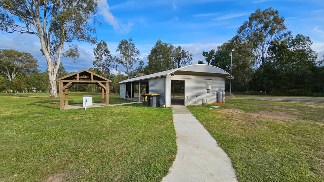 A small, corrugated metal building with a curved roof is situated on a grassy area. A concrete path leads to the entrance. To the left, there is a wooden shelter with a pitched roof and a small white utility box underneath. Trees are scattered in the background, and there are a few bins near the building. The sky is partly cloudy.