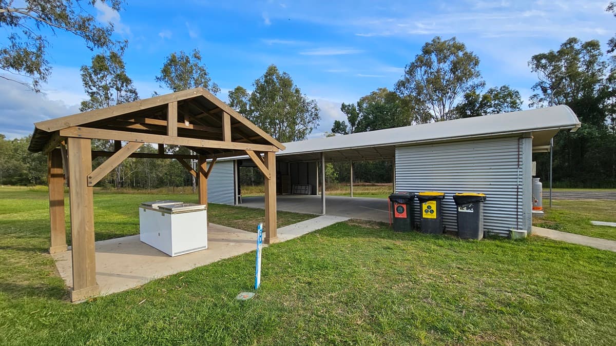 A grassy area with a wooden shelter on the left, housing a white barbecue unit on a concrete slab. To the right, a corrugated metal building with an extended roof provides shade. In front of the building are several wheelie bins in different colours. Trees are visible in the background, and the ground is covered with grass.