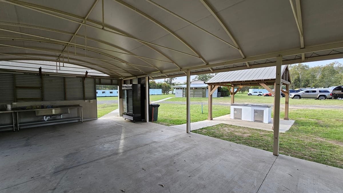 A covered outdoor area with a curved metal roof and concrete floor. On the left, there is a metal sink and counter. In the centre, a large refrigerator stands near an open doorway leading to a grassy area. On the right, a small pavilion with a metal roof and a countertop is visible. In the background, there are several parked cars and buildings, surrounded by trees.