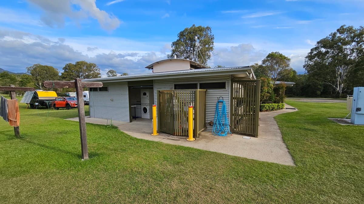 A small, single-storey building with a metal exterior and a flat roof is in the centre. It has an open entrance revealing a washing machine inside. To the right, a blue hose is coiled on the wall. Yellow bollards are in front of a wooden screen. On the left, a clothesline with towels is visible. In the background, there are trees and a few parked vehicles, including a yellow trailer and an orange car. The ground is covered with grass.