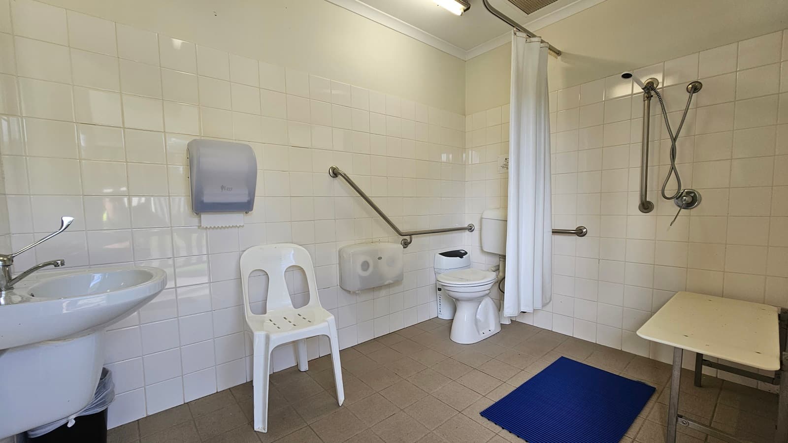 A bathroom with white tiled walls and a beige tiled floor. On the left, there's a sink with a lever tap and a wall-mounted paper towel dispenser above a white plastic chair. In the centre, a toilet with a grab rail beside it. On the right, a shower area with a handheld showerhead and another grab rail, separated by a white curtain. A blue mat is on the floor near the shower, and a small bench is positioned next to it. The ceiling has a fluorescent light fixture.