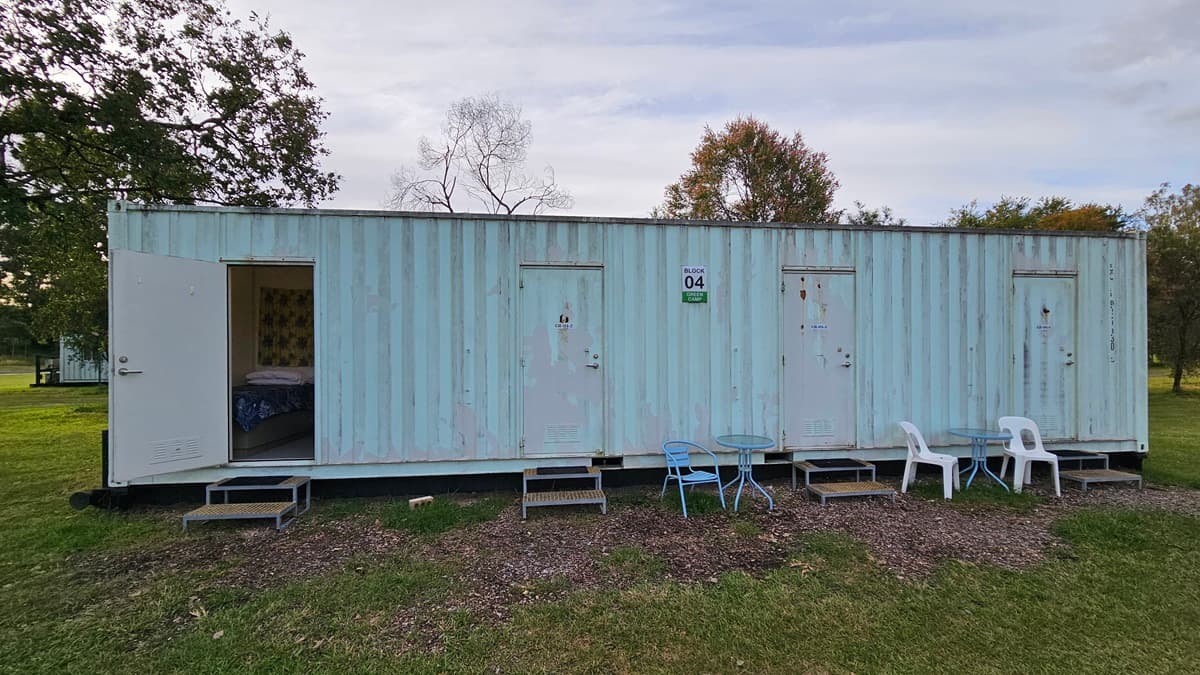 A long, light blue shipping container with four doors, each accessed by small metal steps. The leftmost door is open, revealing a glimpse of the interior. In front of the container, there are two white plastic chairs and a blue metal chair, along with a small table. The container is set on a grassy area with some trees in the background. The ground in front of the container is covered with patches of gravel.