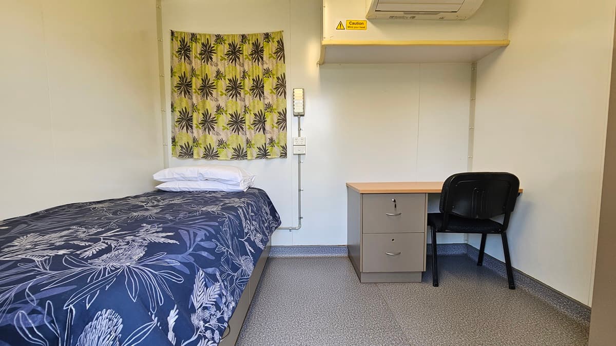 A small room with a single bed on the left, covered with a dark blue floral-patterned duvet and white pillows. Above the bed is a yellow curtain with a black starburst design. On the right, there's a wooden desk with a black chair. Above the desk is an air conditioning unit with a caution sign. The floor is grey and speckled, and the walls are plain white.