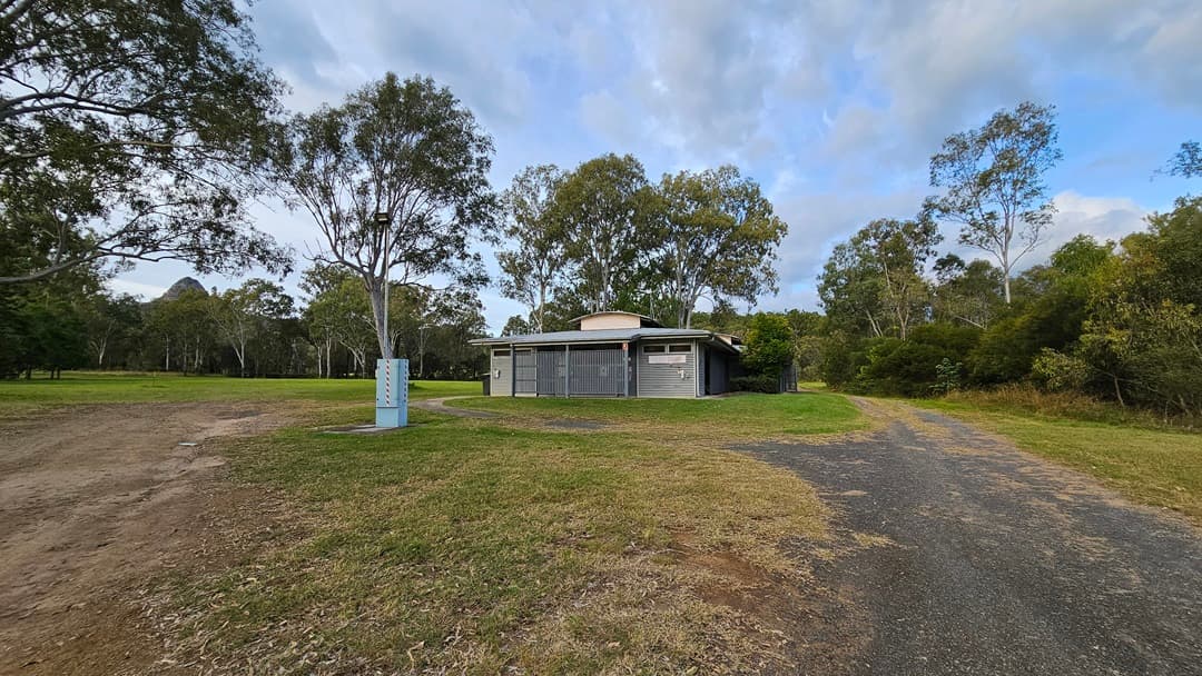 A modern, single-storey building with a flat roof and grey slatted exterior. It is surrounded by a grassy area with a few small bushes in front. On the left and right sides, there are rubbish bins near the building. In the background, there are tall trees with green foliage. The sky is overcast.