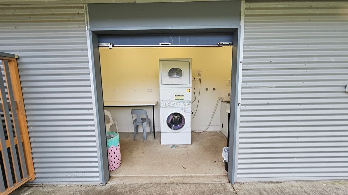 A small laundry room with corrugated metal walls. In the centre, there's a white stacked washer and dryer. To the left, a plastic chair and a laundry basket with a polka dot design. On the right, a countertop with a sink and some plumbing fixtures. The floor is concrete, and the ceiling is not visible.