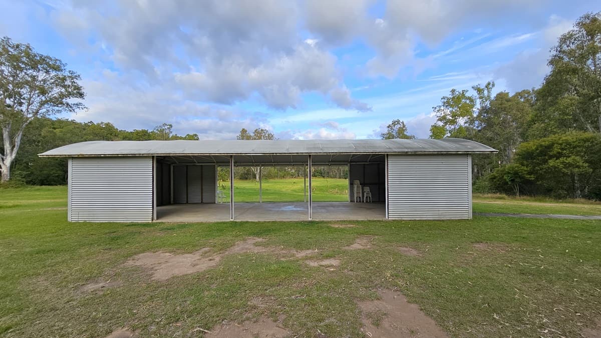A simple, open-sided structure with a corrugated metal roof and walls stands on a grassy area. The building has three open sections supported by metal poles, with the middle section providing a view of the green landscape beyond. Inside, there are a few stools visible on the right. Trees and bushes surround the area, creating a natural backdrop. The ground is a mix of grass and patches of bare earth.