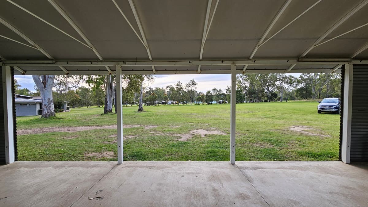 A view from inside a covered area with a concrete floor, looking out onto a grassy field. The ceiling is supported by white beams. In the background, there are several trees scattered across the field. To the right, a parked car is visible on the grass. On the left, a building is partially visible behind the trees.