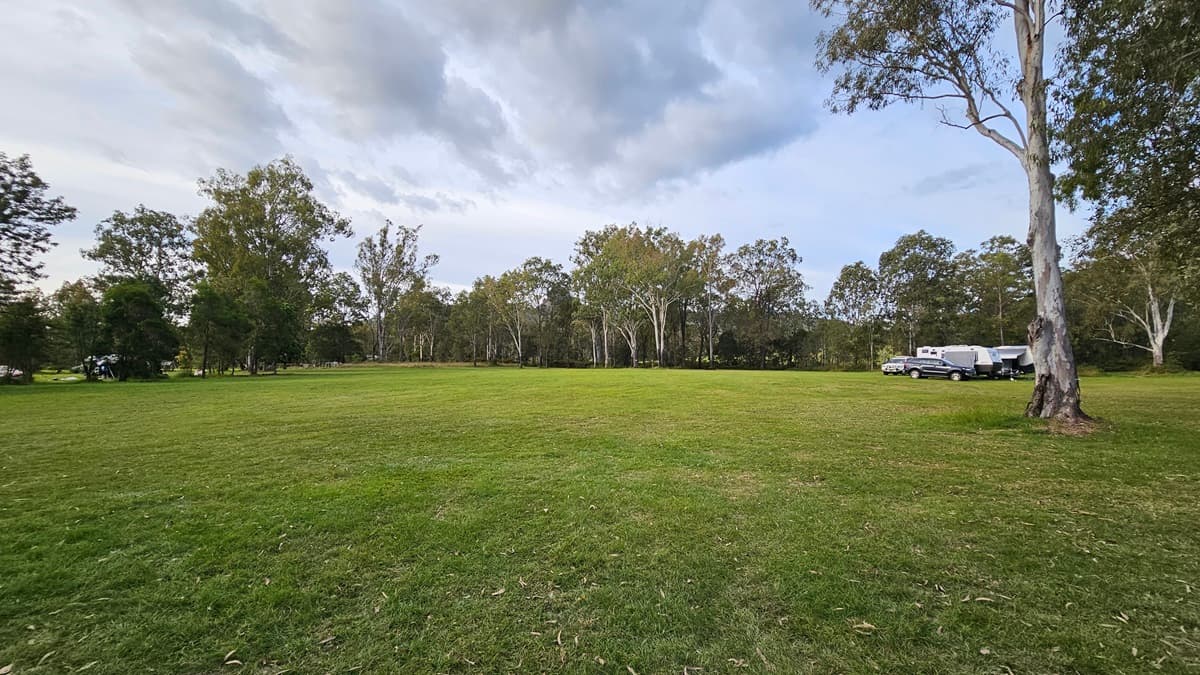 A grassy field stretches across the foreground, with a line of trees in the background. On the right, there are several parked caravans and a car near a large tree. The sky is overcast with grey clouds.