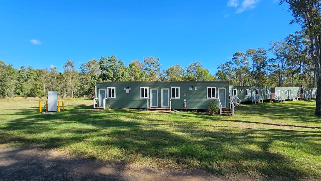 A dirt path runs through a grassy area, leading towards a row of long, green portable buildings in the background. Tall trees with slender trunks are scattered throughout the scene, casting shadows on the ground. The sky is clear and blue, and the area is surrounded by more trees, creating a wooded backdrop.