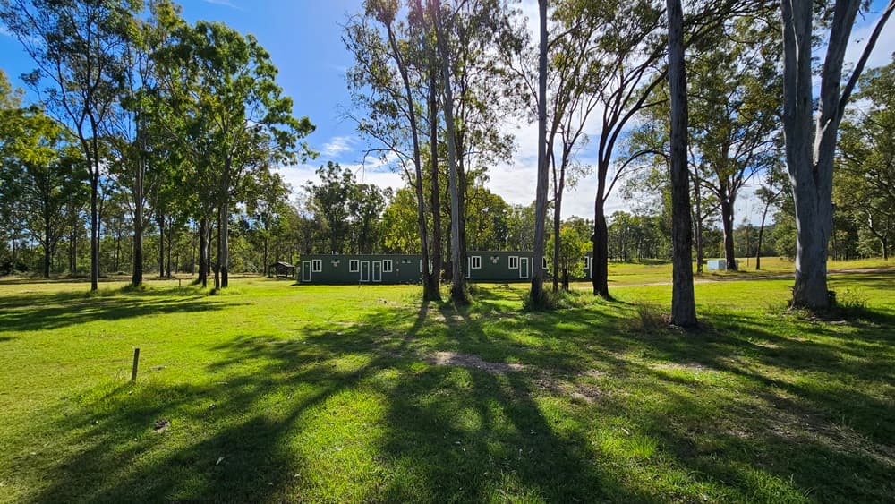 A grassy field with tall trees scattered throughout. In the middle, a long, single-storey building with multiple windows is visible. The shadows of the trees stretch across the grass, indicating sunlight from above. The background is filled with more trees, creating a wooded area. The scene is peaceful and open, with a clear view of the landscape.