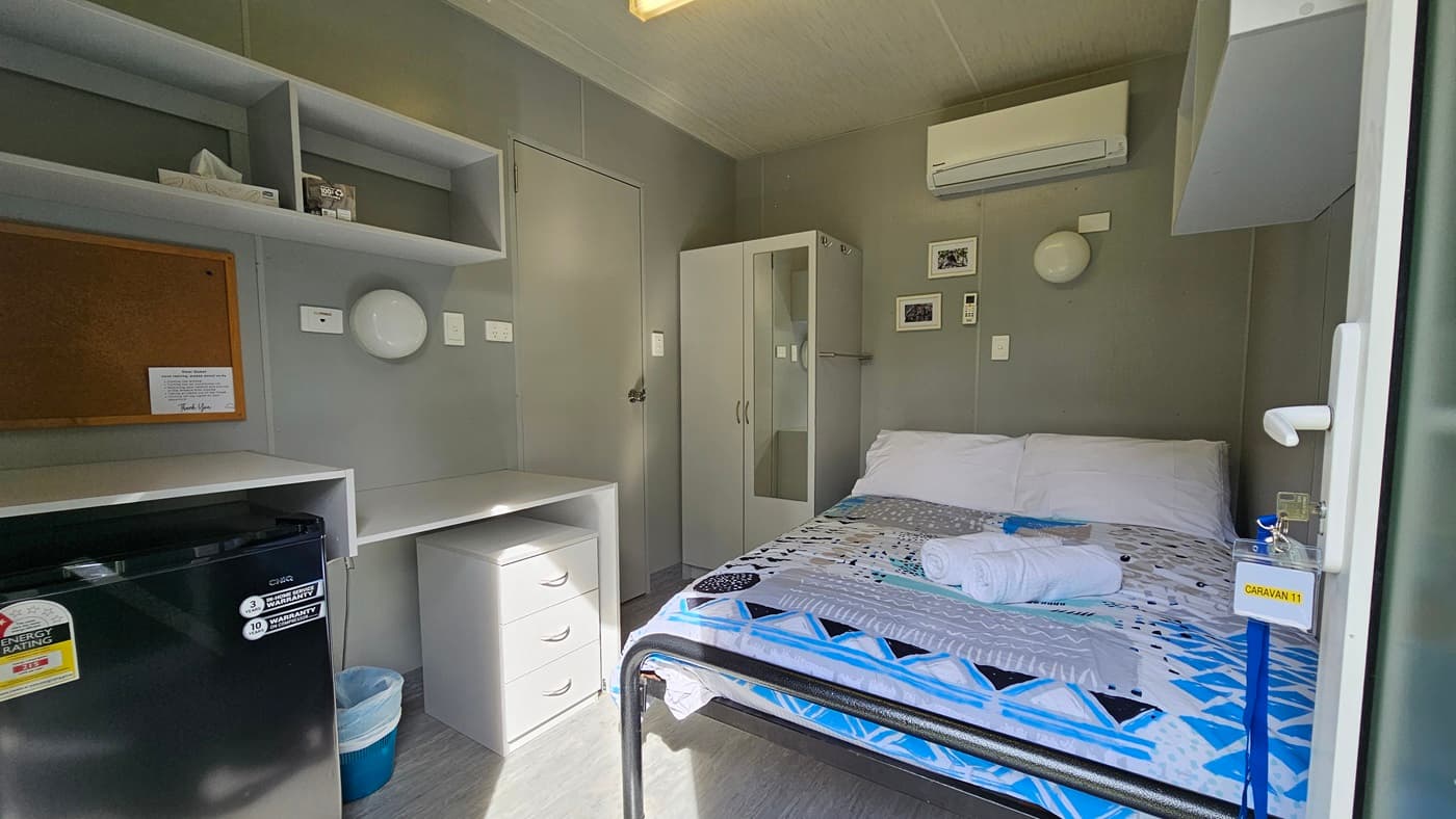 A compact room with a bed on the right, covered with a geometric-patterned bedspread and a towel on top. Above the bed is an air conditioning unit. On the left, there are shelves with a tissue box and a corkboard. Below the shelves is a small fridge and a set of drawers. A wardrobe is next to the bed. The walls are grey, and the floor is light-coloured. The ceiling has a fluorescent light fixture.