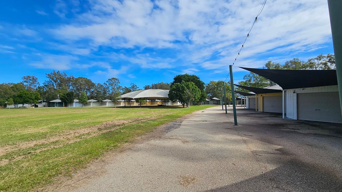 A grassy field on the left with a row of small, round huts in the distance. In the centre, a larger building with a grey roof is partially obscured by trees. On the right, a paved path runs alongside a series of closed stalls with dark shade sails overhead. The background features a line of trees under a partly cloudy sky.