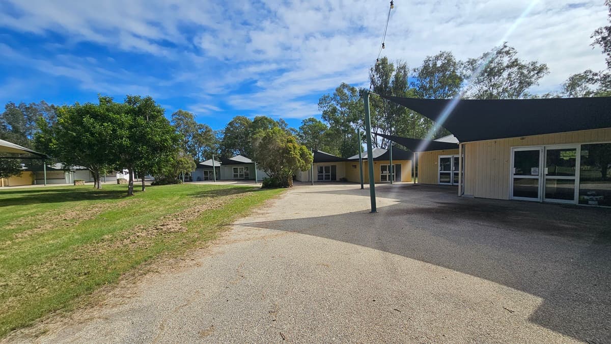 A paved pathway curves through a grassy area with several trees on the left. On the right, there are buildings with large windows and dark triangular shade sails. In the background, more trees and buildings are visible. The sky is partly cloudy.