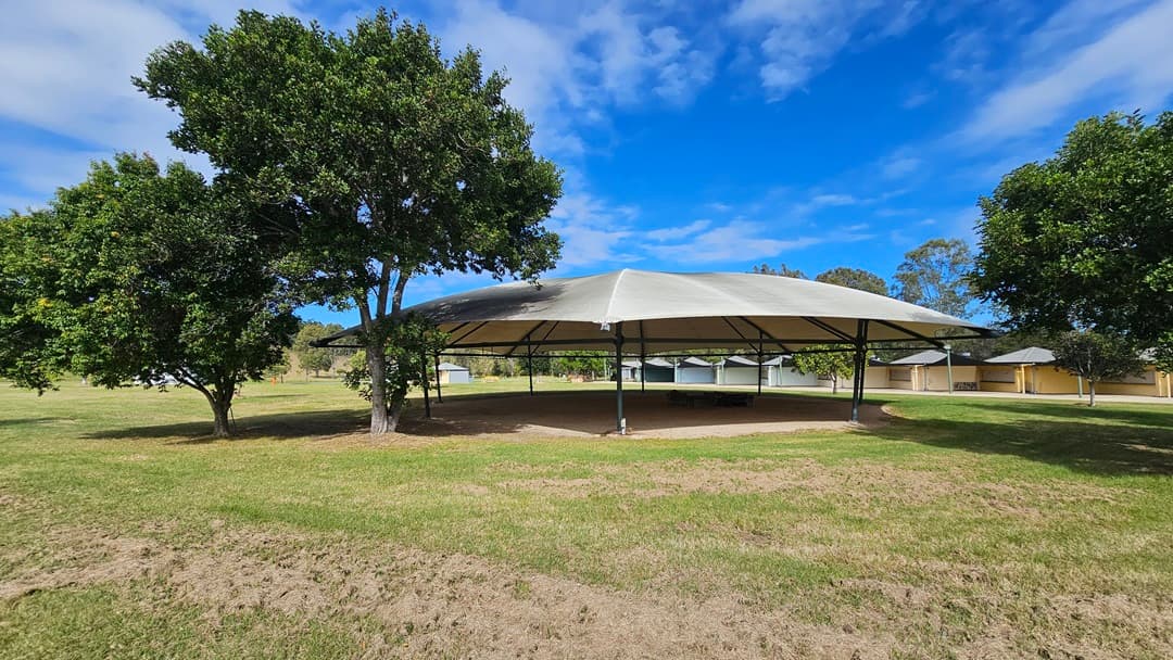 A large, open-sided pavilion with a white, tent-like roof is situated in the centre of a grassy area. On the left and right, there are several green trees providing shade. In the background, there are more pavilions and a few scattered trees. The ground is covered with grass, and the sky is partly cloudy.