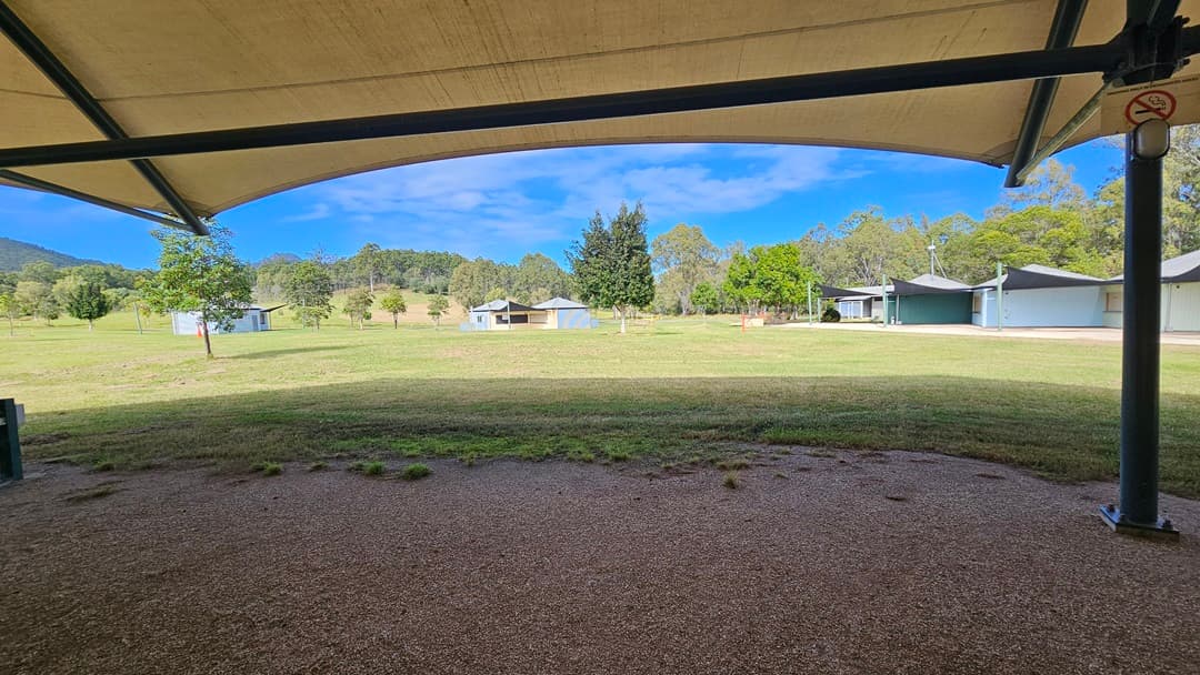A grassy field stretches across the middle of the image, with several small buildings scattered in the distance. Trees are visible in the background, providing a natural border. In the foreground, there is a gravel area under a large, curved canopy supported by metal poles. The canopy provides shade over the gravel, contrasting with the sunlit grass beyond.