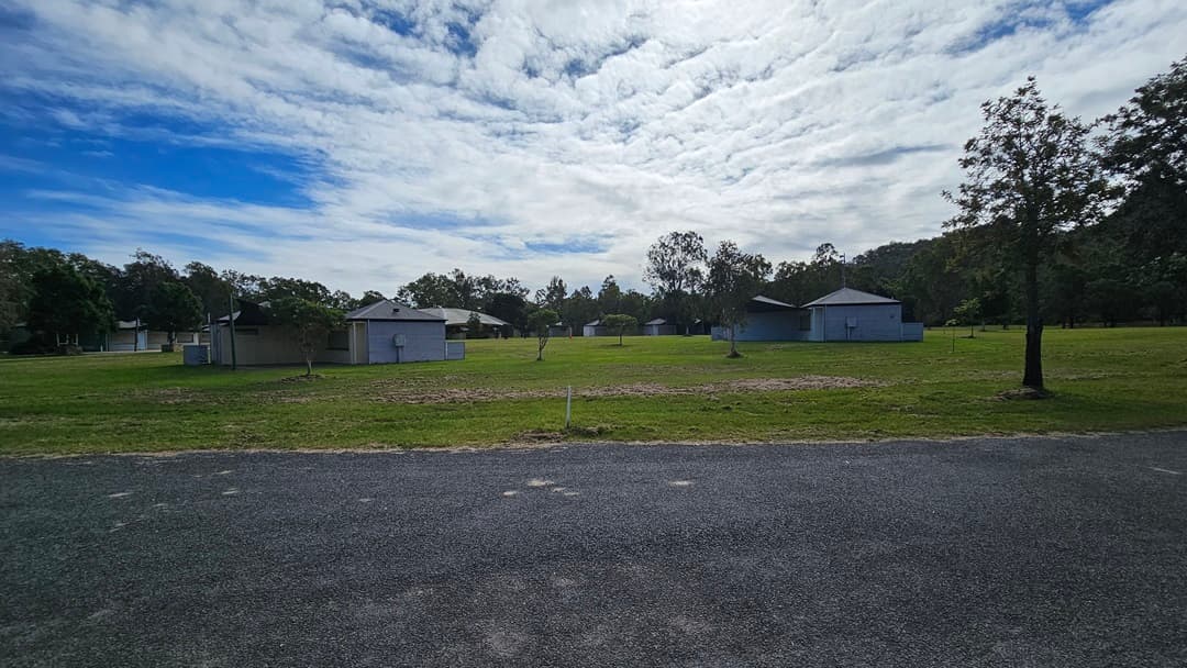 A grassy field with several small, light blue buildings scattered across it. Trees are visible in the background, surrounding the area. In the foreground, there is a gravel path or road. The sky is partly cloudy, casting a soft light over the scene.