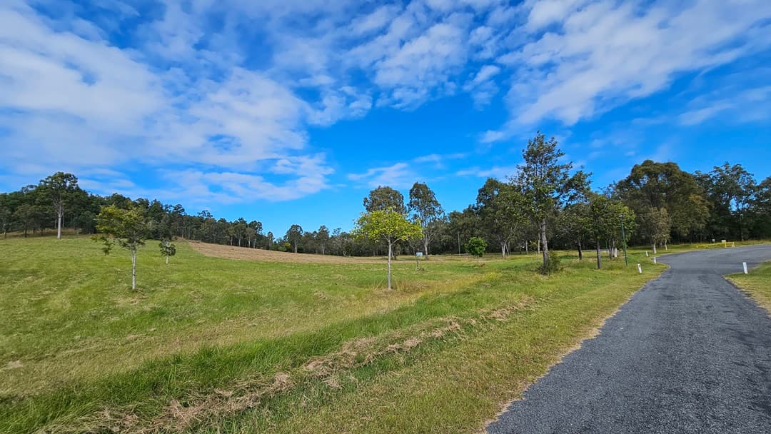 A landscape with a grassy field on the left, dotted with small trees. On the right, a gravel path curves through the scene, bordered by more trees. The background features a line of taller trees along the horizon. The sky is partly cloudy with patches of blue visible.