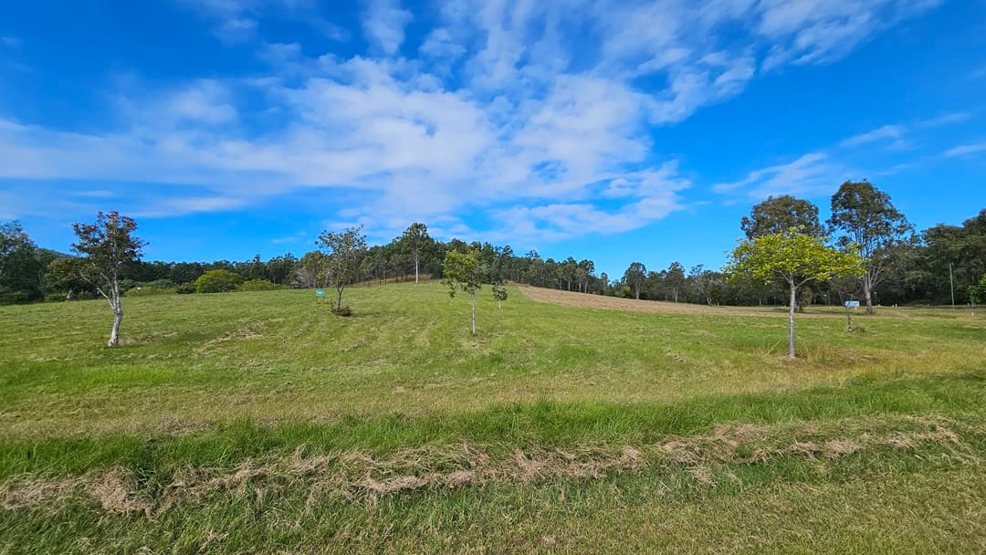 A grassy field with a gentle slope, scattered with small trees. In the background, a line of taller trees marks the edge of the field. The grass is a mix of green and brown, indicating some areas are more dry. The sky is mostly clear with a few clouds.