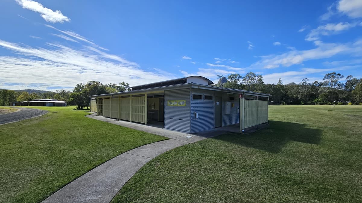 A single-storey building with a corrugated metal exterior is situated on a grassy field. The structure has a flat roof and a covered entrance. A concrete path leads to the building, curving from the foreground. In the background, there are trees and another similar building. The sky is clear with some clouds.
