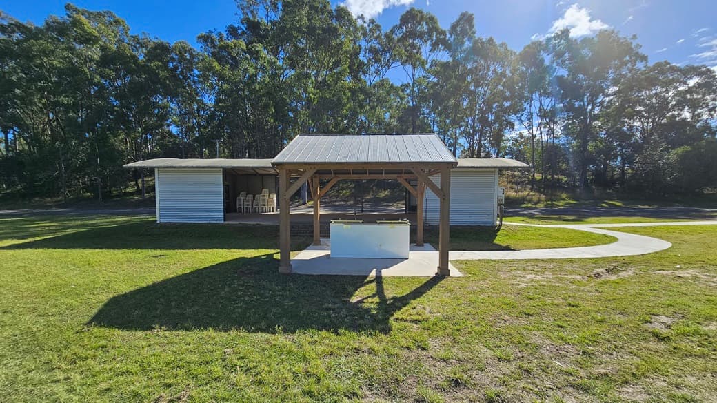 A small wooden shelter with a metal roof stands in the centre of a grassy area. In front of it is a white rectangular structure, possibly a table or bench. Behind the shelter, there is a larger building with a corrugated metal exterior and open sides, containing stacked chairs. A curved concrete path runs from the foreground to the right side of the image. Tall trees with dense foliage form the background.