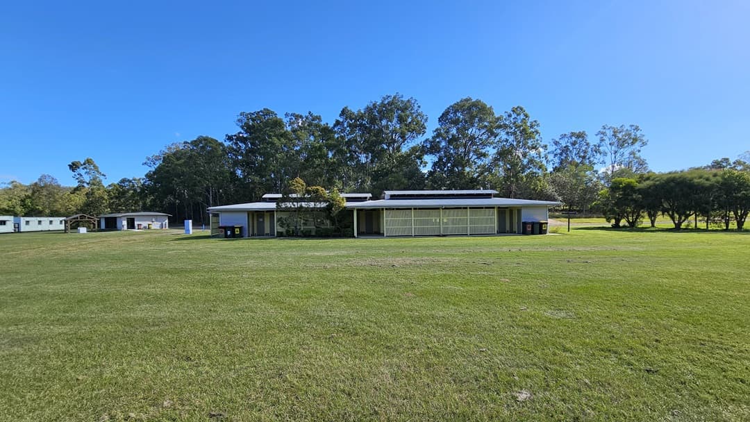 A grassy field with a low, rectangular building in the centre, featuring a flat roof and large windows. The building is surrounded by trees in the background. To the left, there are smaller structures, possibly sheds or cabins. The sky is clear and blue.
