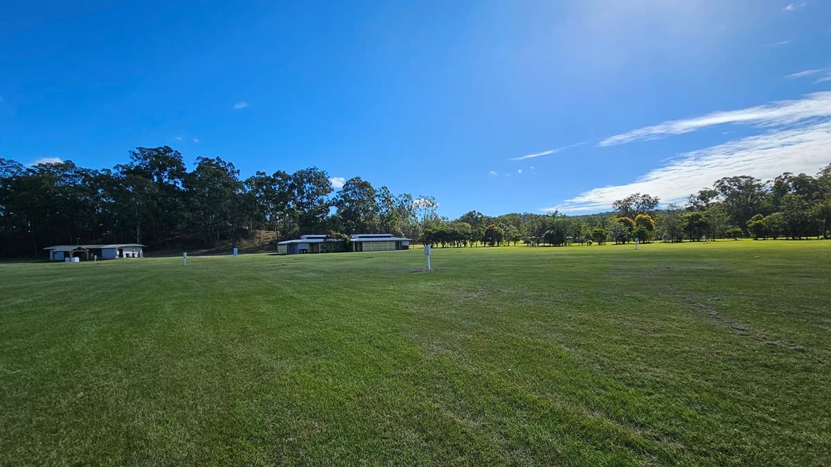 A wide, open grassy field stretches across the foreground, with a few small structures visible in the middle distance. In the background, a line of tall trees forms a dense border. The sky is clear with a few clouds, casting bright light over the scene.