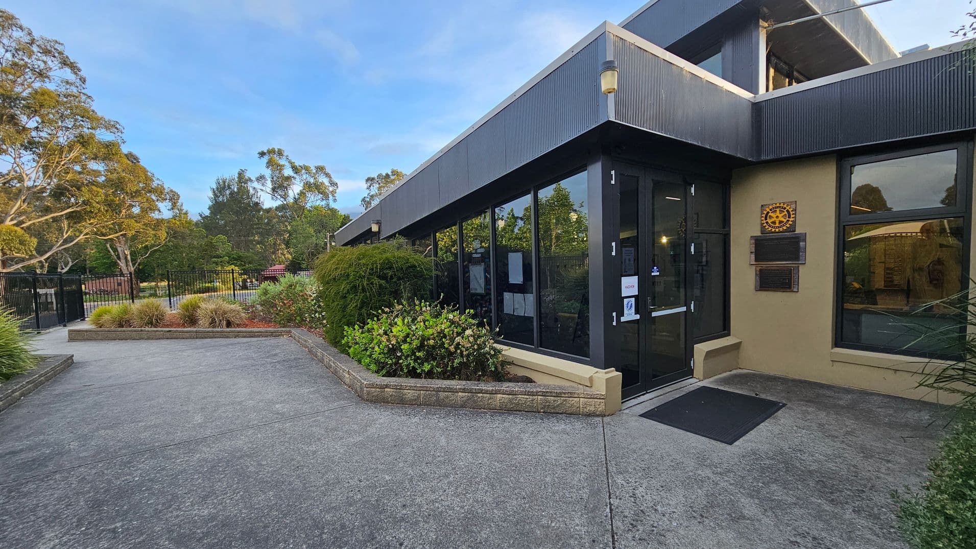 A modern building with large glass windows and a dark exterior is on the right. A plaque with a rotary emblem is mounted on the wall near the entrance. The entrance has a black door with a small ramp leading up to it. On the left, there is a paved pathway bordered by low brick walls and lush greenery, including bushes and small trees. In the background, a black metal fence and more trees are visible, creating a serene, landscaped setting.