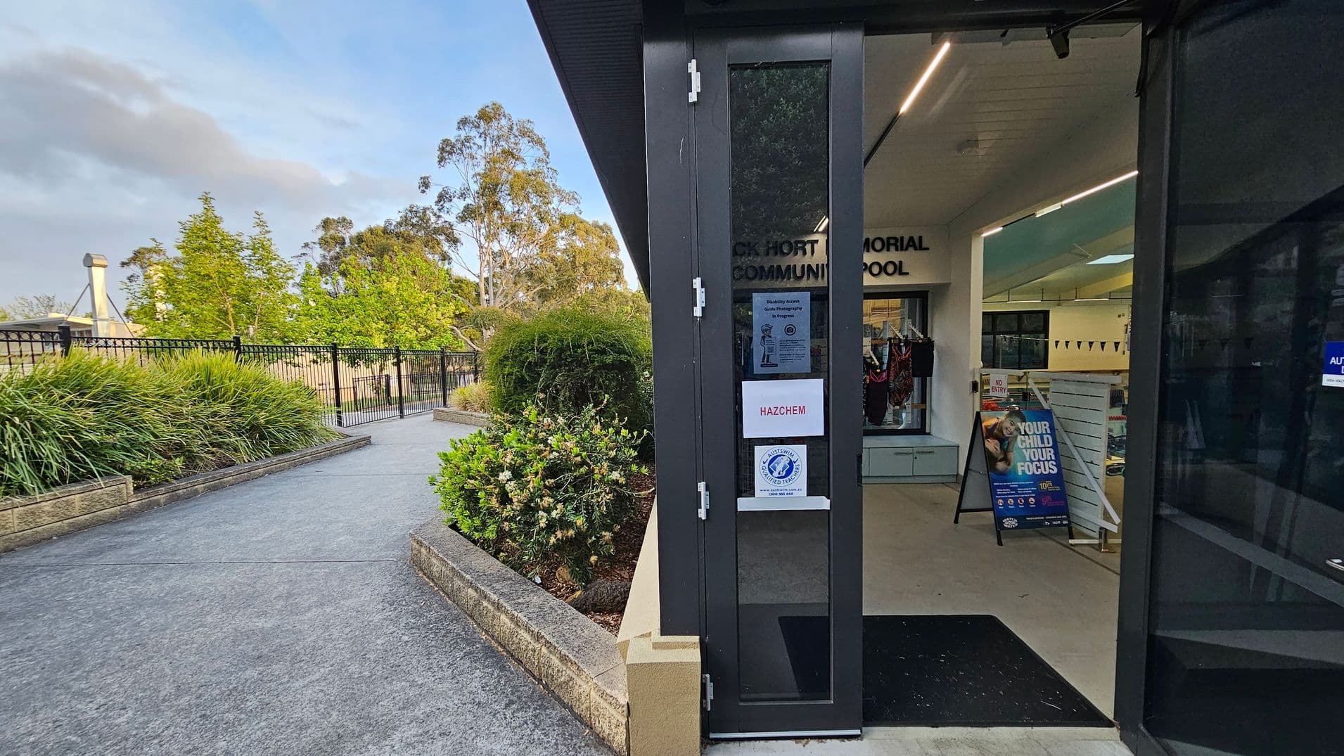 On the left, there's a pathway bordered by lush green bushes and a black metal fence. In the background, tall trees are visible. On the right, a partially open door leads into a building with signs, including one that says "HAZCHEM." Inside, there's a display of swimwear and a poster with the text "YOUR CHILD YOUR FOCUS." The ceiling has recessed lighting, and the floor is light-coloured.