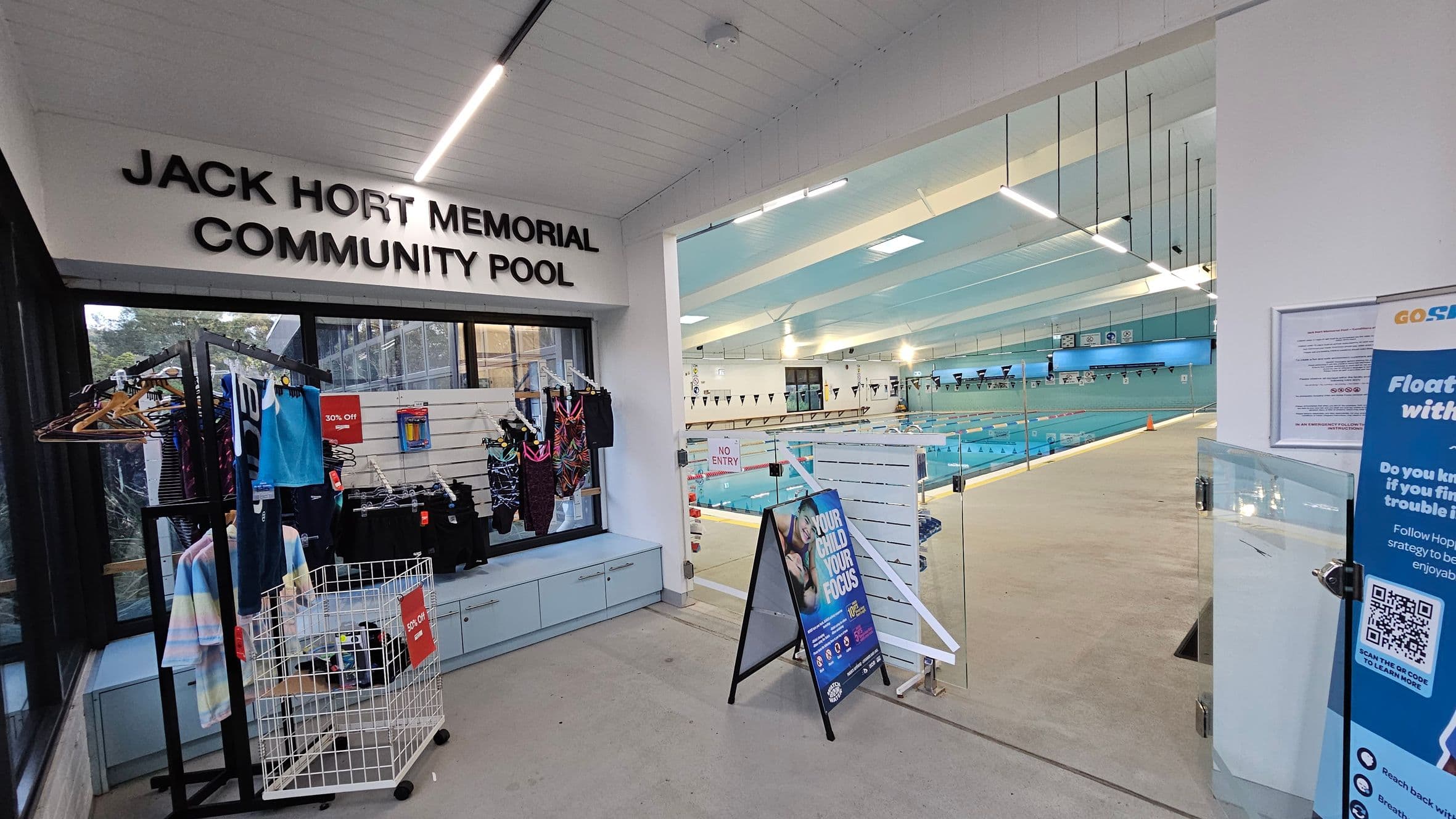 On the left, there is a display of swimwear on hangers and a basket with a "30% Off" sign. Above, the sign reads "Jack Hort Memorial Community Pool." On the right, a glass door leads to an indoor swimming pool with lanes and flags. A stand with a poster and a blue sign with safety information is visible. The ceiling has linear lights, and the floor is light-coloured.