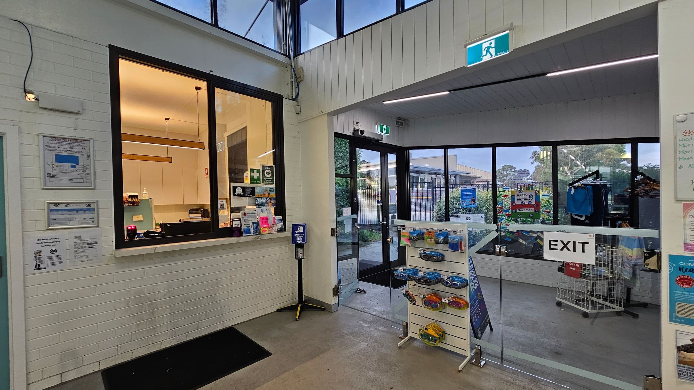 A reception area with a service window on the left, featuring a counter with various items and a lit interior. Notices and signs are displayed on the wall. In the centre, a hand sanitiser dispenser is mounted on a stand. To the right, a glass exit door leads outside, with a rack of clothing and a display stand with packaged items nearby. The ceiling has recessed lighting, and the floor is tiled. An emergency exit sign is visible above the door.