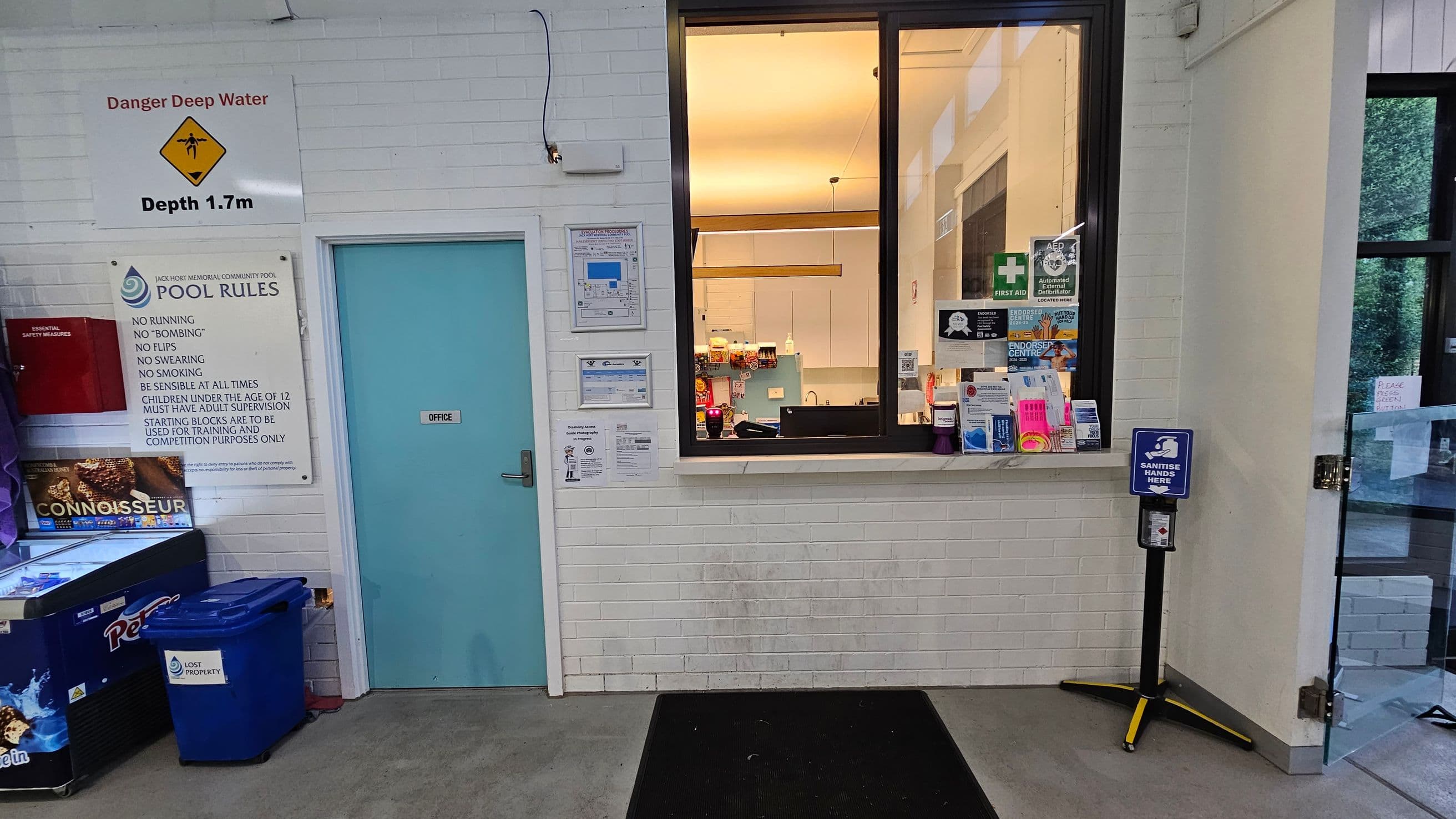 A white brick wall with a blue door labelled "Office" on the left. Above the door, a sign warns of deep water with a depth of 1.7m. Next to it, a board lists pool rules. Below, there's a freezer with "Connoisseur" branding and a blue bin marked "Lost Property". On the right, a window shows an office interior with various notices and a first aid sign. A hand sanitiser station is positioned near a glass door on the far right. The floor is grey with a black mat.
