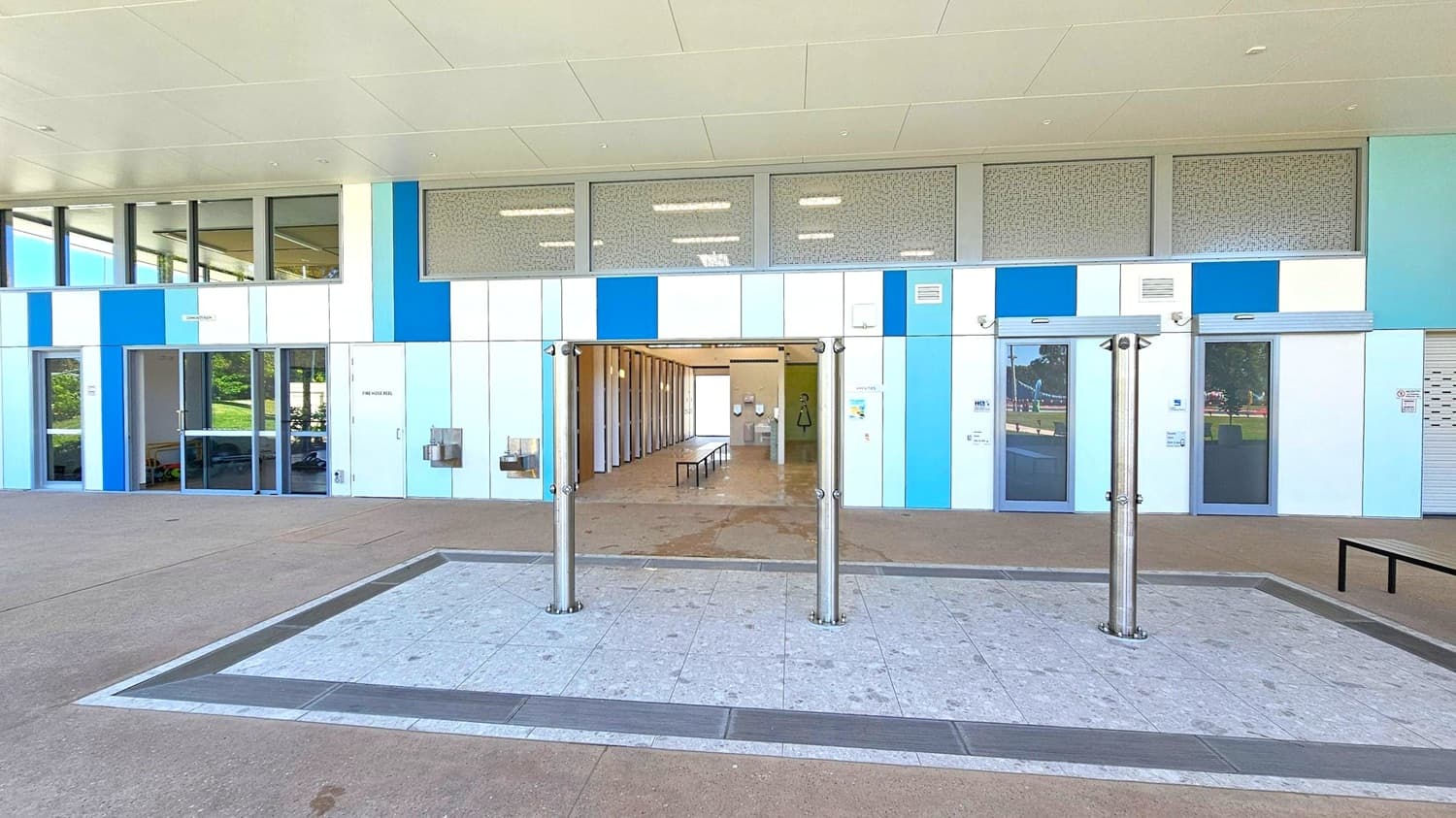 A building facade with a modern design featuring a mix of blue and white panels. In the centre, there is an open entrance leading to an interior space with a bench and several cubicles. Above the entrance, there are large windows with a patterned design. On either side of the entrance, there are closed doors and windows. The ground is paved with a combination of light and dark tiles, and there are three stainless steel bollards in front of the entrance. The ceiling is smooth and white.