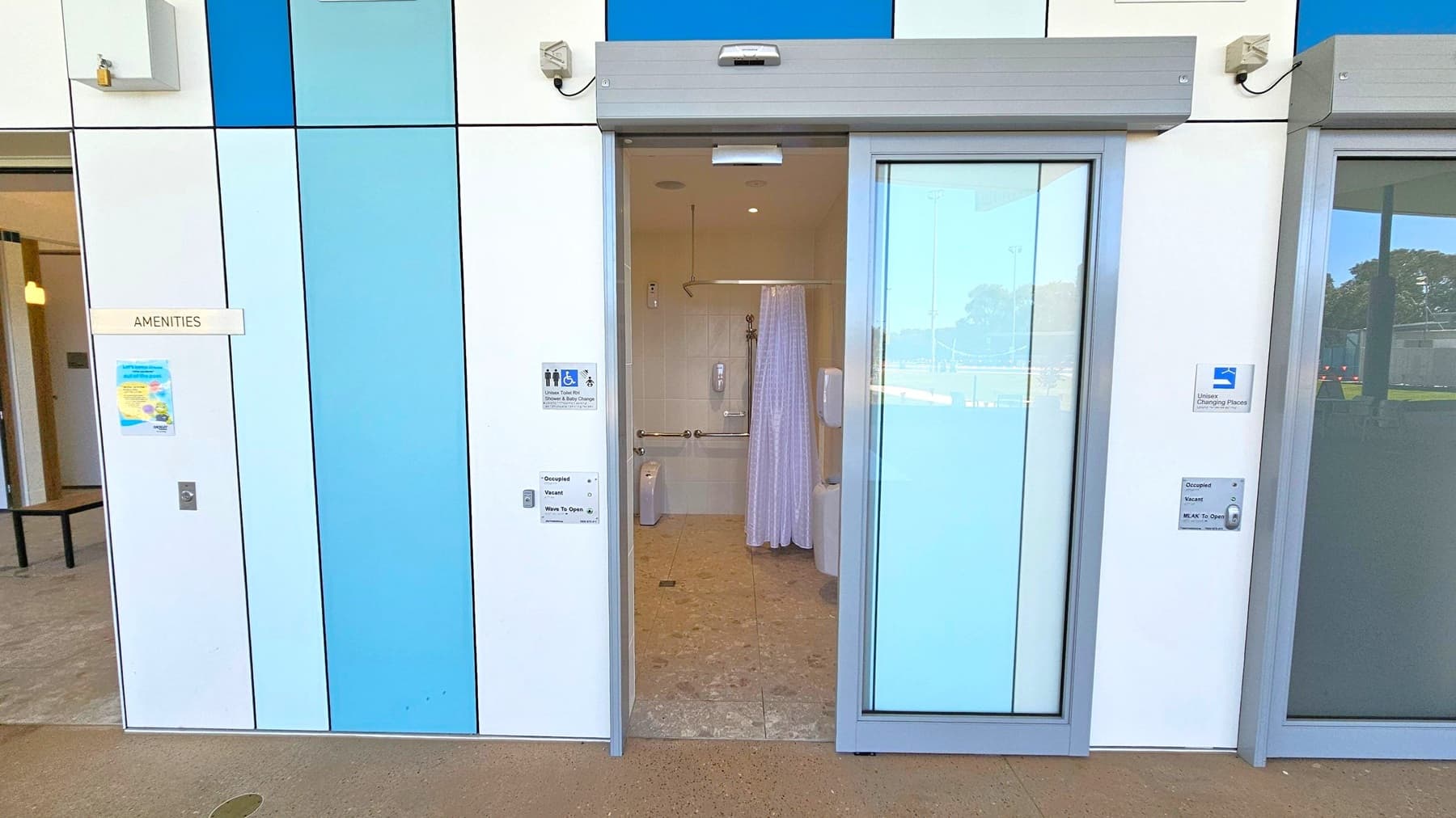 A public amenities facility with a sliding door open, revealing an accessible bathroom inside. The exterior wall is composed of panels in shades of blue and white. A sign on the left reads "AMENITIES" with a poster below it. Inside, there is a toilet, grab bars, and a shower area with a curtain. The floor is tiled, and there are soap dispensers on the wall. The ground outside is concrete, and there are signs indicating occupancy status and usage instructions.
