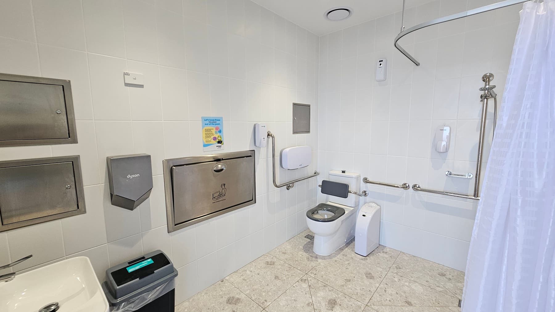 A public restroom with white tiled walls and a light-coloured tiled floor. On the left, there is a sink with a waste bin below, a Dyson hand dryer, and two metal panels. In the centre, a baby changing station is mounted on the wall. On the right, there is a toilet with support rails, a wall-mounted soap dispenser, and a shower area with a curtain. The ceiling has a vent and a small sensor.