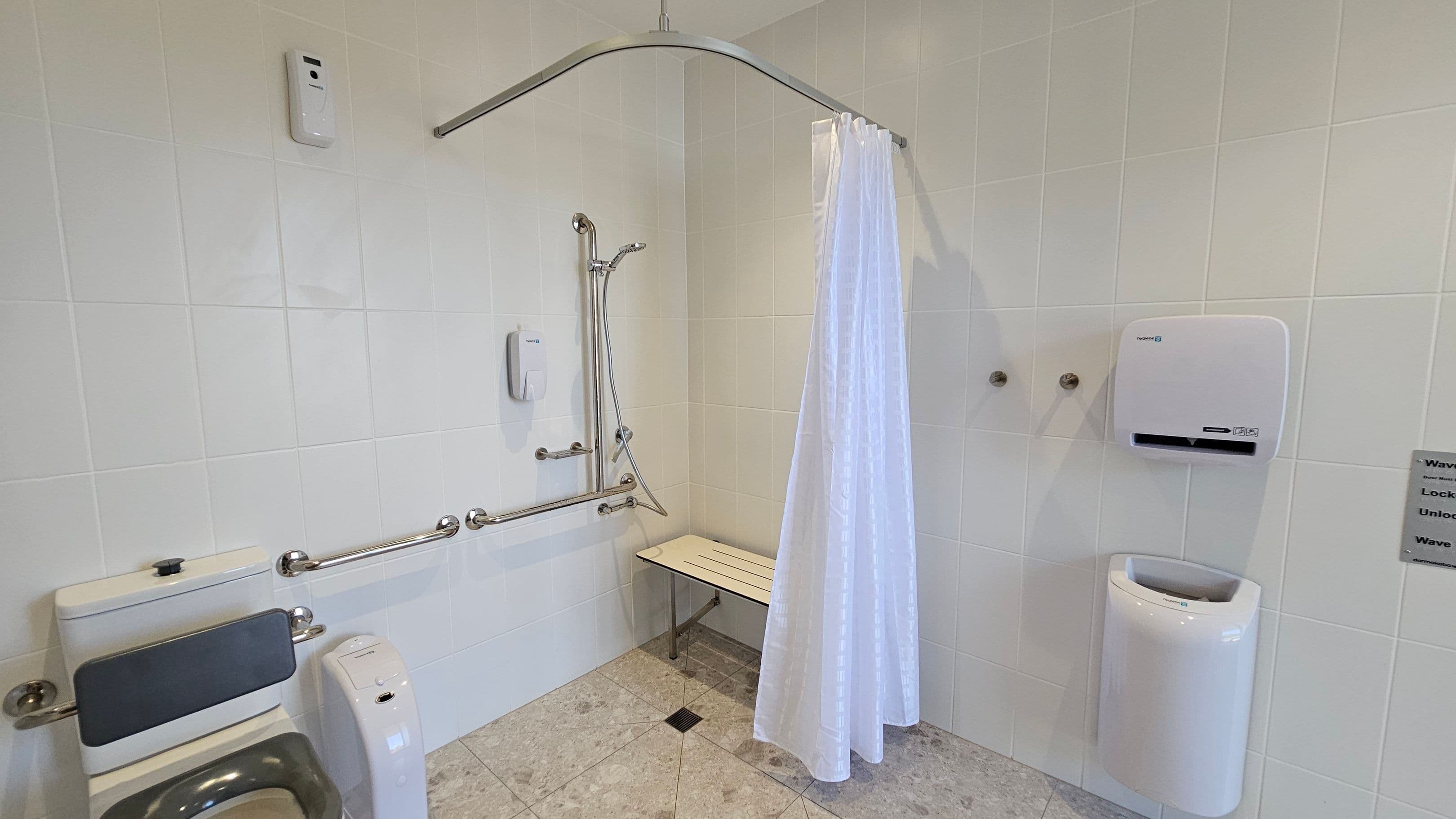 A bathroom with white tiled walls and a light-coloured tiled floor. On the left, there is a toilet with a backrest and grab bars. In the centre, a shower area features a handheld showerhead, grab bars, and a foldable bench. A white shower curtain hangs from a curved rod. On the right, a wall-mounted hand dryer and a paper towel dispenser are visible. A small sign with instructions is also present on the right wall.
