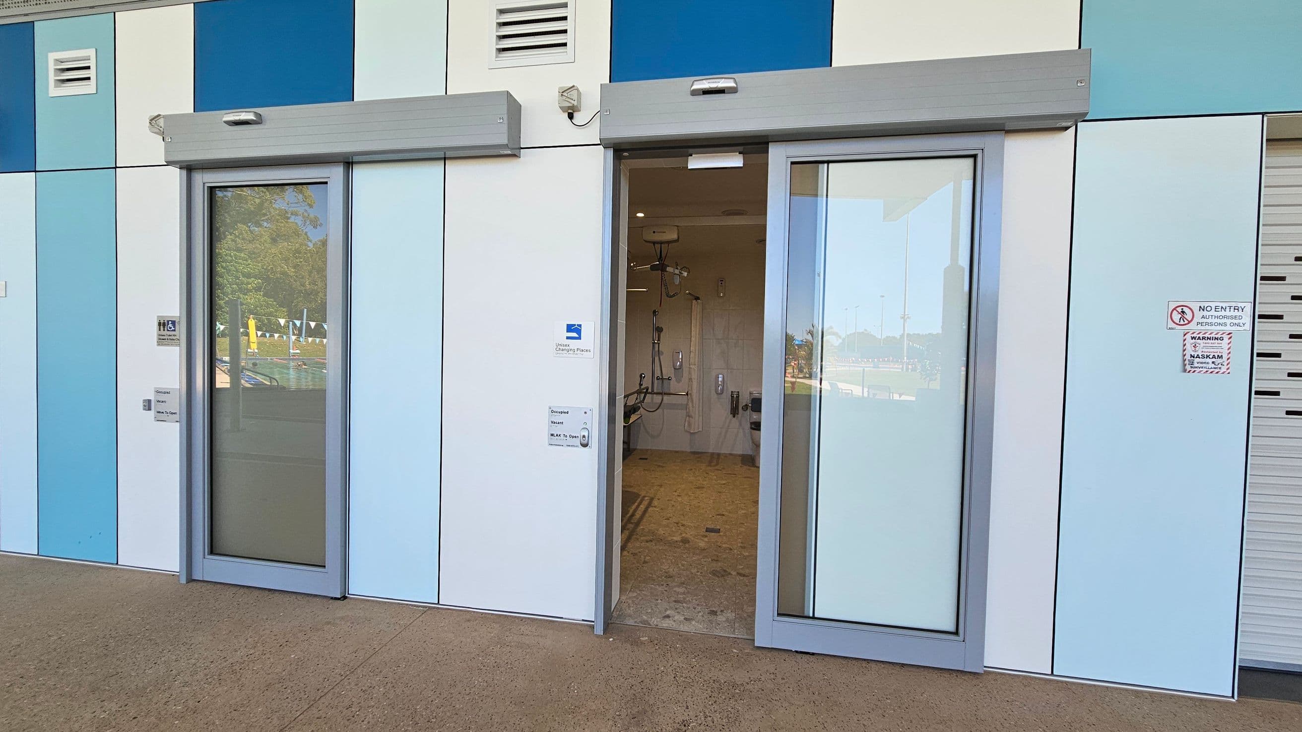 A building exterior with a series of vertical panels in shades of blue and white. Two sliding glass doors are present; the left door is closed, while the right door is partially open, revealing an interior with shower facilities. Above each door are grey metal awnings. Signs near the doors indicate accessibility features and entry restrictions. The ground is paved with concrete, and there is a reflection of a pool area in the glass.
