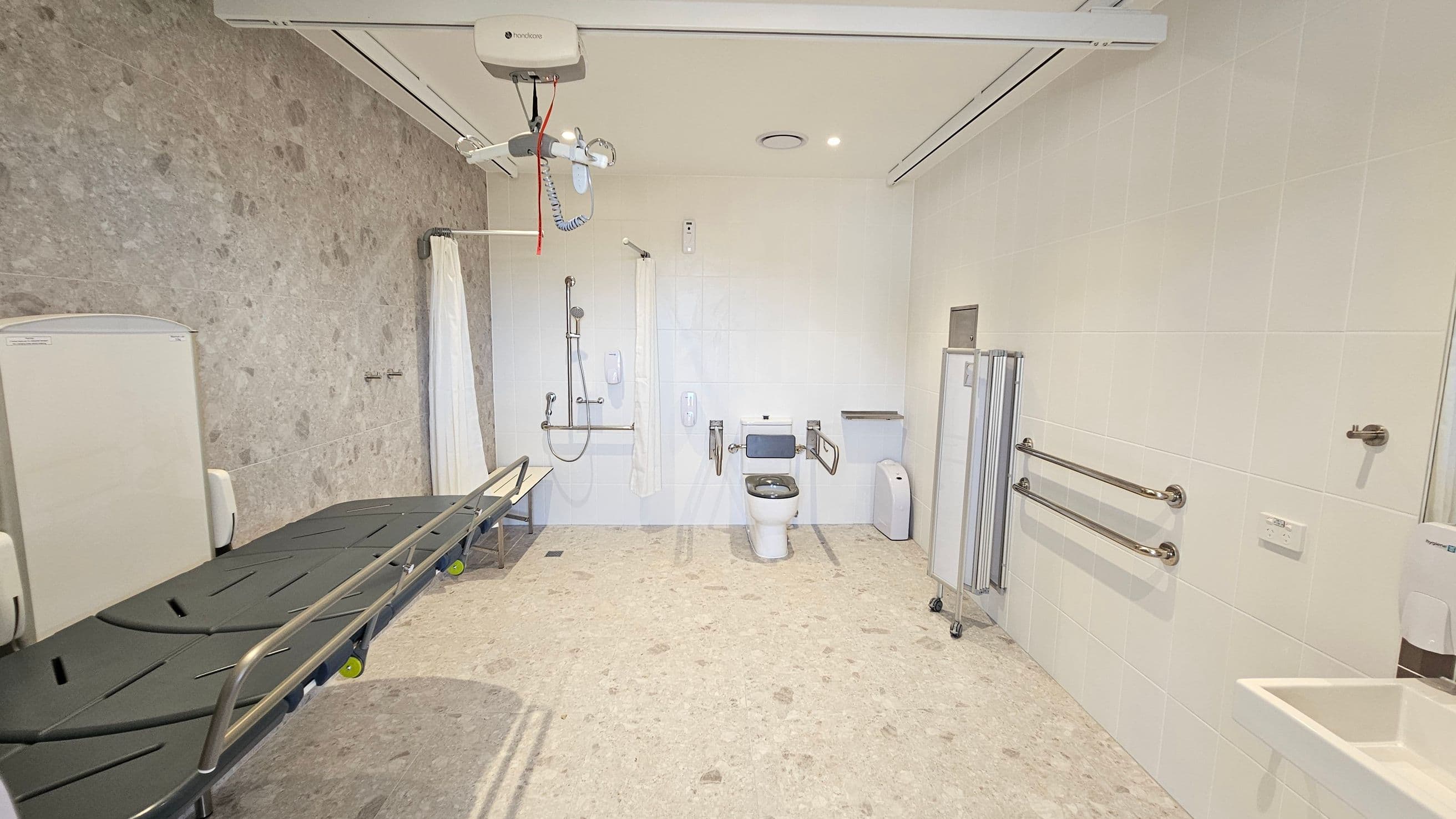 A spacious, accessible bathroom with a beige tiled floor and light-coloured walls. On the left, a large, adjustable changing table with side rails is positioned against the wall. In the centre, a toilet with support bars on either side is visible, along with a shower area equipped with a curtain and handrails. On the right, there are additional support bars and a small sink. Ceiling-mounted equipment for assistance is present, and a privacy screen is folded nearby.