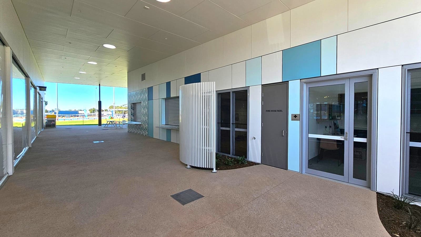 A covered outdoor area with a tiled ceiling featuring circular lights. The right side has a wall with a mix of blue and grey panels, a door marked "Fire Hose Reel," and a glass door with a medical cross sign. A curved white slatted structure is in front of the wall. The left side has large glass windows, and the floor is a mix of concrete and textured surfaces. In the background, there is a view of a grassy area with a picnic table and a distant structure.