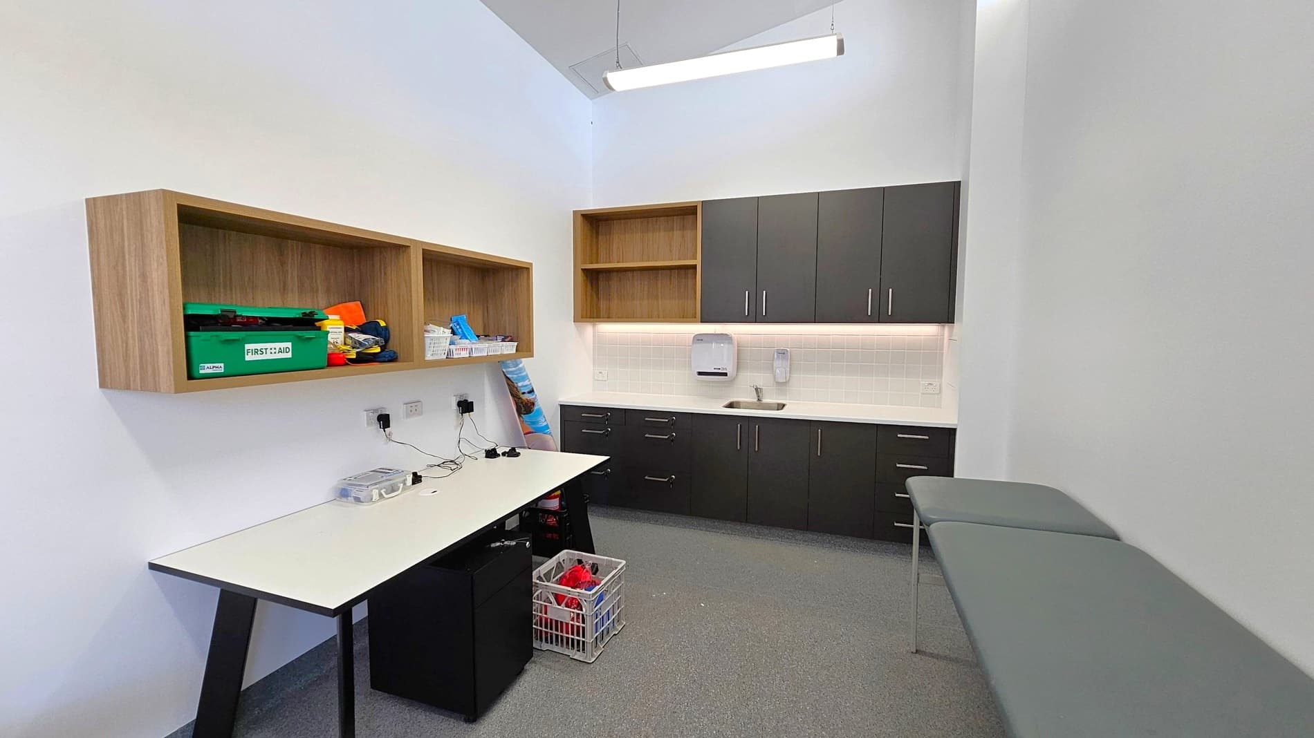 A small room with a grey carpeted floor and white walls. On the left, there is a white table with a black frame, above which are wooden shelves containing a green first aid kit and other items. In the centre, black cabinets with silver handles are mounted on the wall, with a small sink and a paper towel dispenser below. On the right, there are two grey examination tables. A fluorescent light fixture hangs from the ceiling.
